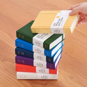 Könnte beinhalten: Ein Stapel aus sechs 2027 Prism 365 Daily Diary Planern in Gelb, Dunkelgrün, Hellblau, Marineblau, Lila und Rot, jeweils mit einer weißen Papierbanderole. Der oberste gelbe Planer wird angehoben und gibt den Blick auf die weißen Seiten frei.