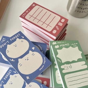 May include: A stack of rectangular paper memo pads in red, blue, and green. The red pad features a to-do list with cat icons. The blue pad is labeled DON'T FORGET and shows two cat faces. The green pad is labeled MEMO with a speech bubble and cat art.