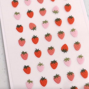 May include: A sheet of strawberry stickers. The stickers feature red and pink strawberries with green tops. Some strawberries are solid red, while others are pink with white polka dots. The stickers are on a white background.