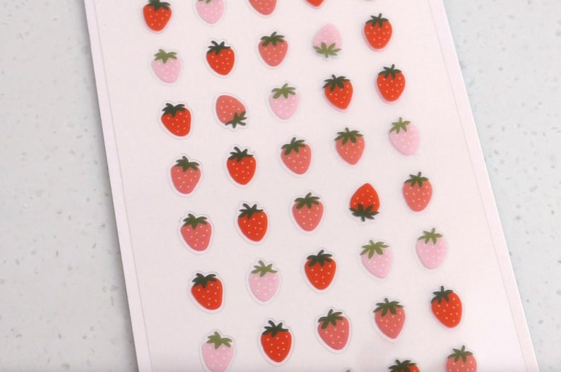 May include: A sheet of strawberry stickers. The stickers feature red and pink strawberries with green tops. Some strawberries are solid red, while others are pink with white polka dots. The stickers are on a white background.