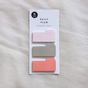 Può includere: Un biglietto bianco con la scritta "DAILY PLAN" e tre blocchetti adesivi rosa, grigi e corallo. Il blocco superiore ha un motivo a righe diagonali. I blocchi hanno la forma di linguette per cartelle.
