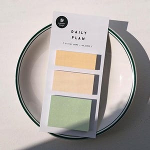 May include: A white card with the text "DAILY PLAN" and three sticky notes in yellow, beige, and green. The card rests on a white plate with a green rim.
