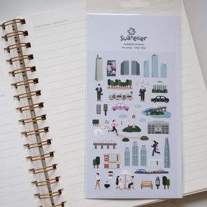 May include: A sheet of stickers featuring illustrations of buildings, people, and cars in a city setting. The stickers are on a white background with the text "Suatelier" and "Suatelier stickers no. 223 city city!" printed in black.