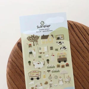 Puede incluir: Una hoja de pegatinas Suatelier con ilustraciones en miniatura de una casa, vacas, un puesto de frutas y otras escenas rurales. Las pegatinas tienen un fondo verde claro y azul con el texto "Suatelier" y "good afternoon".