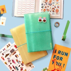 May include: A collection of stationery items, including a teal glittery journal with a birthday cake sticker, a yellow journal, stickers, washi tape, and a notebook with labels. The journal is secured with a teal elastic band. The image is on a light blue background.
