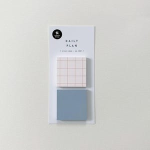 May include: A white card with the text "DAILY PLAN" and two sticky notes. The top sticky note is white with a pink grid pattern, and the bottom one is a solid light blue. The card also has a small black logo.