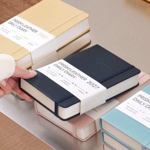 May include: A collection of Prism Leather 2027 daily diaries in various colors including yellow, navy blue, pink, and light blue. Each diary features a white paper band with the text Prism Leather 2027 Daily Diary, 365 days, 416 pages, and Indigo.