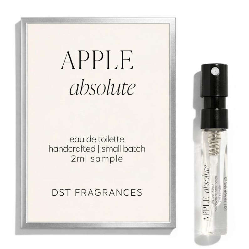 May include: A rectangular card with a silver border featuring the text APPLE absolute, eau de toilette, handcrafted | small batch, 2ml sample, and DST FRAGRANCES, alongside a small clear glass spray vial containing liquid with a black cap and label.