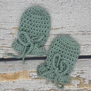 May include: A pair of crocheted baby mittens in a light green colour. The mittens have a tie at the wrist.