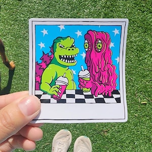Puede incluir: Un monstruo verde con dientes afilados y un monstruo rosa con ojos viscosos están sentados en una mesa a cuadros, cada uno sosteniendo un batido. La imagen está enmarcada en un marco blanco estilo Polaroid con fondo azul y estrellas blancas.