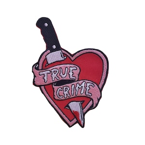 May include: A red heart with a black knife sticking through it. The knife has a pink ribbon wrapped around it with the words "TRUE CRIME" written on it.