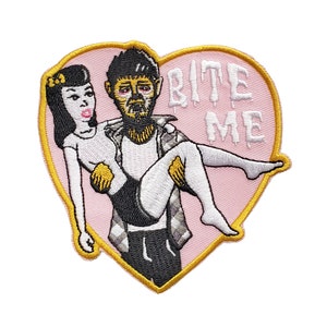 Puede incluir: Parche bordado termoadhesivo en forma de corazón rosa con contorno dorado. El parche presenta una imagen de dibujos animados de un hombre cargando a una mujer con el texto "BITE ME" en letras blancas.