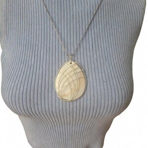 May include: A teardrop-shaped pendant featuring a smooth, cream-colored shell inlay encased in a delicate silver-toned wire cage. The pendant hangs from a thin, matching silver-toned chain, resting against a light blue ribbed fabric surface.