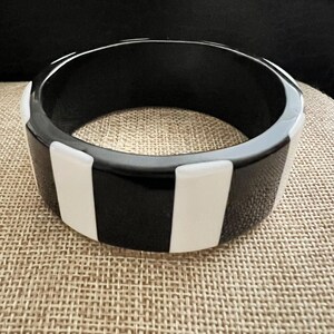 May include: A wide, circular bangle bracelet featuring a high-contrast pattern of alternating vertical black and white rectangular segments. The bracelet has a smooth, polished finish and a thick, solid band construction with a uniform cylindrical shape.
