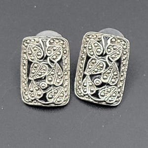 May include: A pair of silver-toned rectangular stud earrings. Each earring features a detailed leaf and dot design within a rectangular frame. The earrings have a post backing for secure wear.