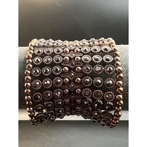 May include: Wide cuff bracelet with a dark bronze finish. The bracelet features rows of small, dark-colored gemstones set in a floral pattern, with a beaded edge. The center has a decorative, geometric design.