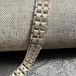 May include: Close-up of a silver-toned bracelet with a repeating rectangular link design. The bracelet is made of multiple rows of connected links, creating a textured appearance. The background is a gray, textured fabric.