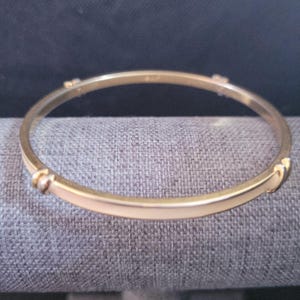 May include: A thin, circular gold-tone bangle bracelet featuring a smooth, polished finish. The metal band is accented with small, evenly spaced decorative nodes or segments along its circumference, creating a simple and continuous circular design.