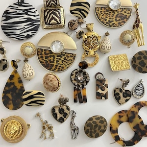 May include: A collection of vintage animal print jewelry, including earrings, pendants, and brooches. The pieces feature zebra, leopard, and tortoise shell patterns, with gold and rhinestone accents. The assortment includes giraffe and lion motifs.