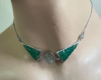 Puede incluir: Un collar de tono plateado que presenta dos segmentos triangulares de piedra verde tallada a los lados de un colgante cuadrado de filigrana. La cadena está compuesta por delicados eslabones plateados con pequeños detalles florales circulares.