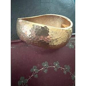 May include: A gold-toned cuff bracelet with a hammered texture. The wide, open design features a smooth interior and a textured exterior. The bracelet is displayed on a dark surface with a floral design.