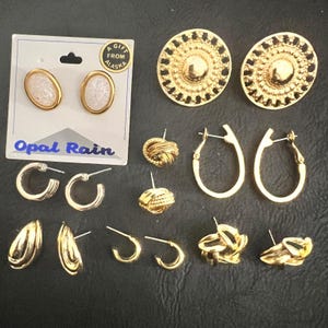 May include: A collection of gold-toned earrings, including hoop, stud, and clip-on styles. Some earrings feature textured designs, while others have a smooth, polished finish. One pair is on a card that says "Opal Rain" and "A Gift From Alaska".