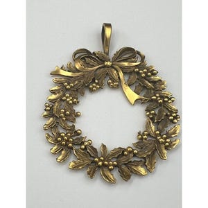 May include: A gold-colored wreath pendant with a bow at the top. The wreath is made of leaves and berries, and it has a loop for hanging. The pendant is likely made of metal and is designed for decorative purposes.