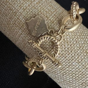 May include: A gold-toned metal chain bracelet featuring a toggle clasp with a twisted rope texture. A small, shield-shaped charm hangs from the clasp, clearly engraved with the brand name FORNASH in capital letters.