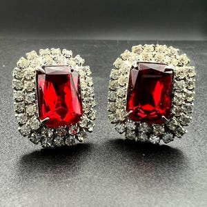 May include: A pair of vintage earrings featuring large, rectangular, ruby-red stones surrounded by a halo of small, clear crystals. The earrings have a silver-tone metal setting and a classic, elegant design.