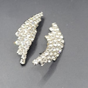 May include: A pair of silver-toned, wing-shaped earrings. Each earring is encrusted with numerous small, sparkling crystals, creating a shimmering effect. The earrings are displayed against a dark gray background.