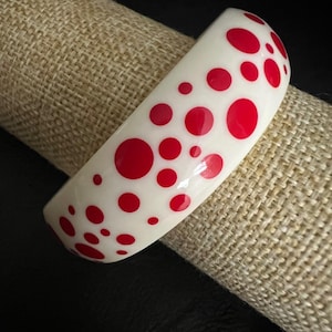 May include: A smooth, off-white bangle bracelet featuring a scattered pattern of vibrant red circular dots of varying sizes across its curved surface. The bracelet has a glossy finish and a wide, rounded profile that maintains a consistent thickness.