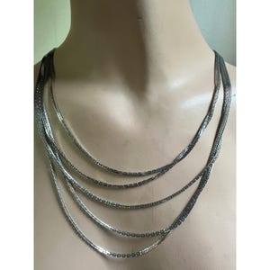 May include: A multi-strand silver-toned necklace. The necklace features five strands of varying lengths, cascading down. The strands are made of small, rectangular links, creating a delicate and elegant appearance. The necklace is displayed on a mannequin.