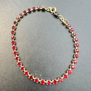 May include: A gold-toned bracelet featuring a series of vibrant red gemstones. The bracelet is composed of small, round, red stones set closely together, creating a continuous line of color. The clasp is also gold-toned.