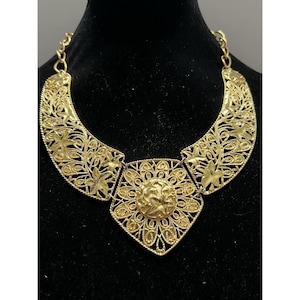 May include: A gold-toned statement necklace with intricate filigree and floral designs. The necklace features a central pendant with a raised floral motif and two curved side pieces, all connected by a delicate chain.