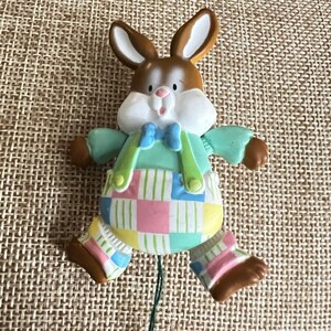 May include: A brown and white rabbit figurine wearing a light blue bow tie, green suspenders, and pastel-colored checkered overalls with pink, yellow, and blue squares. The rabbit has brown ears, a white muzzle, and brown feet with striped socks.