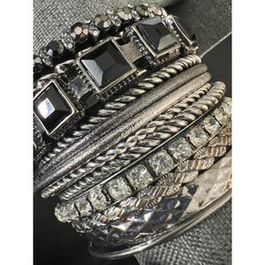May include: A multi-layered bracelet set featuring a combination of metallic bands. The stack includes rows of square black faceted stones, clear crystal accents, twisted silver-tone metal ropes, and textured bands with a polished finish.