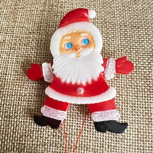 May include: A Santa Claus figurine ornament featuring a red suit with white trim, a white beard, blue eyes, and black boots. The figure has outstretched arms with red mittens and a red hat with a white pom-pom, designed for hanging with a thin red string.