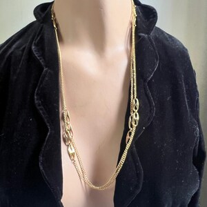 May include: A long, gold-colored necklace with a double chain design. The necklace features a series of oval-shaped links interspersed along the chain. The necklace is displayed against a black velvet jacket.