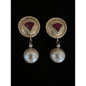 May include: A pair of gold-toned earrings with a circular design, featuring a red, fan-shaped accent. Each earring has a large, dangling, off-white pearl. The earrings are set against a black background.
