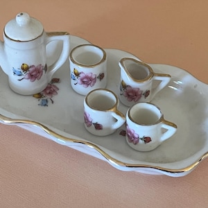 May include: A white porcelain tea set with floral designs. The set includes a teapot, creamer, and four cups. The set is on a white tray with a gold trim.