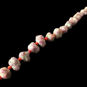 May include: A strand of raw, irregularly shaped ruby beads in shades of pink, red, and green, strung together on a vibrant orange cord. The beads have a rough, natural texture and are arranged in a linear sequence against a solid black background.