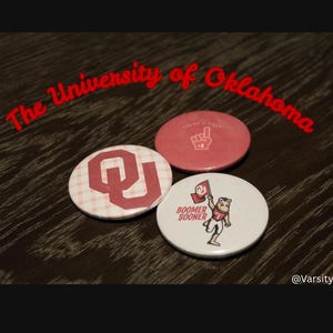 May include: Three circular pin-back buttons featuring University of Oklahoma branding. One button has a red OU logo on a white plaid background, one is solid red with a foam finger graphic, and one is white with a mascot holding a flag and text Boomer Sooner.