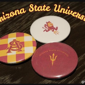 May include: Three circular pin-back badges featuring Arizona State University branding. One badge has a yellow and maroon checkerboard pattern with an AS logo, one is white with a maroon mascot, and one is maroon with a gold trident and text.