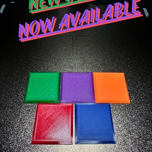 Puede incluir: Cinco posavasos cuadrados en diferentes colores: verde, morado, naranja, rojo y azul. Los posavasos est&aacute;n hechos de un material texturizado y tienen un borde ligeramente elevado. El texto "NEW COLORS NOW AVAILABLE" est&aacute; escrito en letras rojas y verdes sobre un fondo negro.