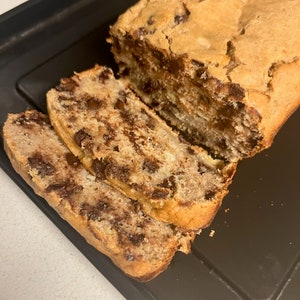 May include: A loaf of banana bread with chocolate chips, sliced and ready to eat. The bread is on a black baking sheet.