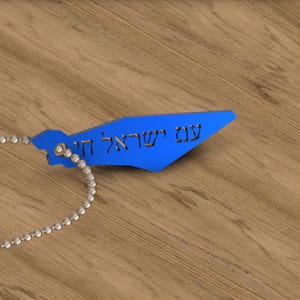 Könnte beinhalten: Ein blauer Anhänger in Form einer Karte mit hebräischen Text, befestigt an einer silbernen Kugelkette. Der Anhänger hat eine leuchtend blaue Farbe und scheint aus Metall oder Kunststoff zu sein. Der Text ist in einer stilisierten Schriftart.