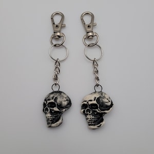 Może przedstawiać: Dwa breloki do kluczy z zawieszkami w kształcie czaszki. Każdy brelok ma srebrny karabińczyk, mały pierścień i łańcuszek prowadzący do zawieszki w kształcie czaszki. Zawieszki w kształcie czaszki są czarno-białe, ze szczegółowymi rysami twarzy.