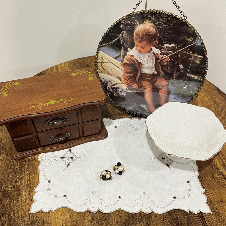 Pu&ograve; includere: Un portagioie in legno con due cassetti e decorazioni floreali &egrave; posto accanto a un piatto decorativo con una scena di un bambino che pesca. Un piatto da portata bianco e un paio di orecchini di perle e neri sono esposti su un runner bianco ricamato.