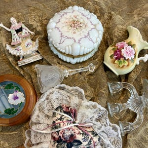 Pu&ograve; includere: Una collezione di oggetti decorativi vintage, tra cui una statuina in porcellana, un portagioie bianco e floreale, una brocca di vetro con rose, un'opera d'arte floreale incorniciata, uno specchio a mano in vetro, un sacchetto floreale e candelabri in vetro.