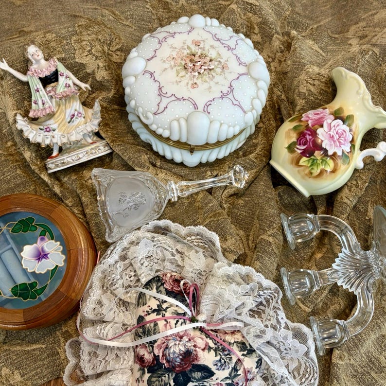 Pu&ograve; includere: Una collezione di oggetti decorativi vintage, tra cui una statuina in porcellana, un portagioie bianco e floreale, una brocca di vetro con rose, un'opera d'arte floreale incorniciata, uno specchio a mano in vetro, un sacchetto floreale e candelabri in vetro.